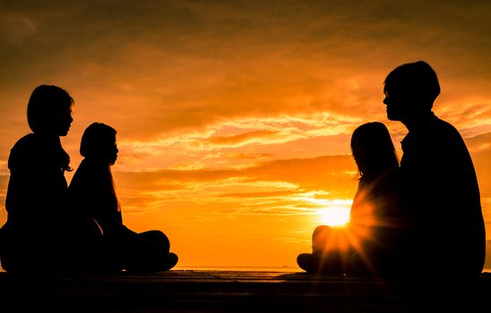 Four young people sit on wooden pier at sunrise on the beach to make meditation with orange  beautiful sky and clouds.