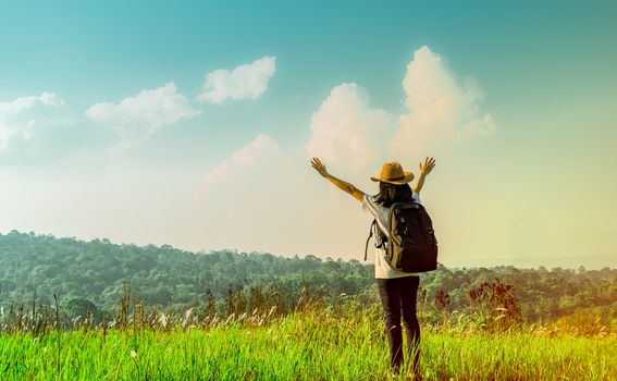 Happy woman tourist with hat and backpack standing and raise her hands up on the hill with green grass field on sunshine day with blue sky and cumulus clouds. Young traveller enjoy beautiful scenery.