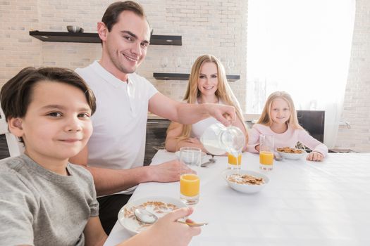 Happy young parents and their children sit together at kitchen table, eat flakes, have healthy breakfast, enjoy good morning, have friendly relationship. Family and eating concept.