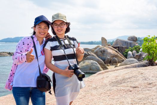 Mother and daughter standing thumb up and smiling happily at Hin Ta Hin Yai and beautiful nature landscape of blue sea and sky during summer travel at Koh Samui island, Surat Thani, Thailand