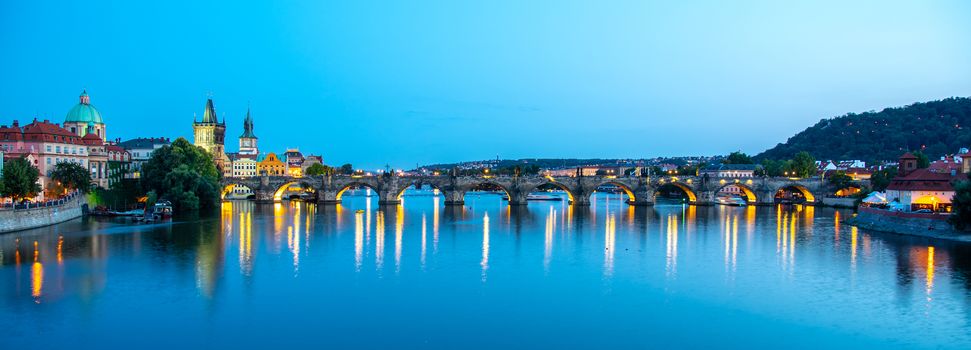 Illuminated Charles Bridge reflected in Vltava River. Evening panorama of Prague, Czech Republic. Panoramic shot.