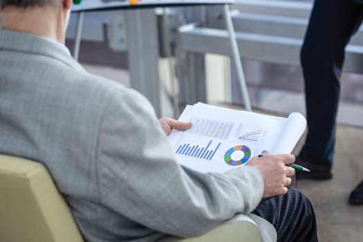 Senior business man sitting at conference, listening to presentation and holding documents with financial diagrams and data