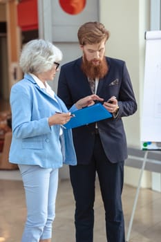 Young business man and senior woman look together at mobile phone and documents