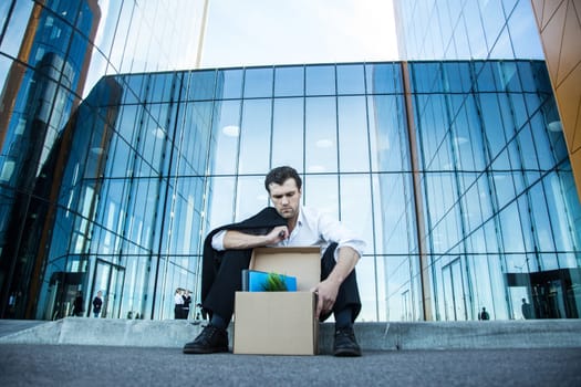 Fired business man sitting frustrated and upset on the street near office building with box of his belongings. He lost work