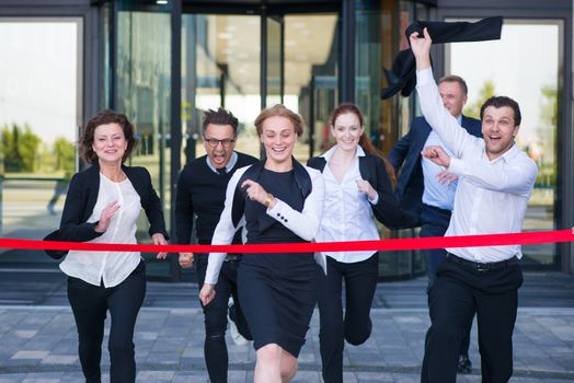 Group of happy business people running from office building crossing red ribbon finish line