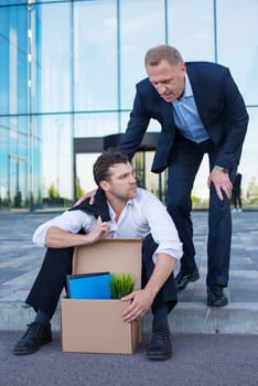 Fired business man sitting frustrated and upset on the street near office building with box of his belongings. He lost work. Other businessman comforts and encourages him