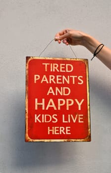 Woman hand holding red metal painted sign, tired parents and happy kids live here, over concrete wall with copy space