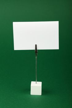 Blank white cardboard sign with copy space on metal note holder over green, paper background, front side view