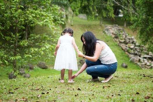 Mother helps little girl wearing shoe at green park. Asian family outdoors portrait.