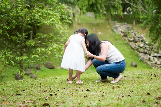 Mother helps little girl wearing shoe at park. Asian family outdoors portrait.