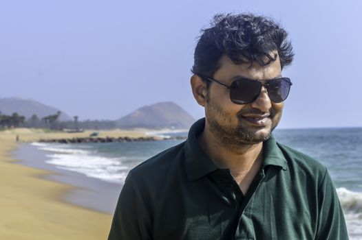 Portrait of elegant Indian man in formal t shirt and sunglasses outdoors, sea backgrounds with copy space.