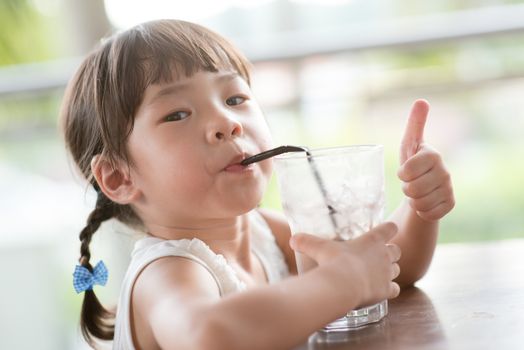 Little Asian girl drinking ice chocolate and giving thumb up at cafe. Natural light outdoor lifestyle.