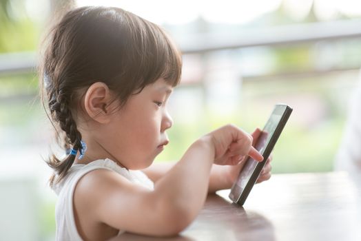 Little Asian girl playing smart phone at cafe. Natural light outdoor lifestyle.