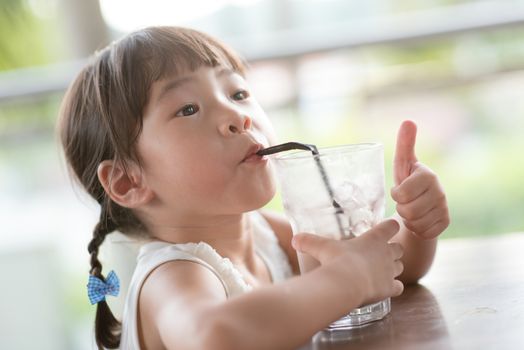 Little Asian child drinking ice chocolate and giving thumb up at cafe. Natural light outdoor lifestyle.