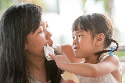 Child wipes mouth for her mom at cafe. Asian family outdoor lifestyle with natural light.