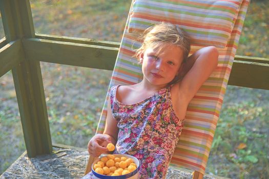 The little girl is sitting on a garden deckchair on a verandah. Small girl is eating cheese flavoured snacks in the garden. Dreamy and romantic image. Summer and happy childhood concept.