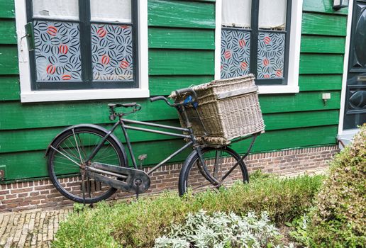 old bicycle with a worn wicker basket in front in front of old wooden house from green painted wood and red and black curtains