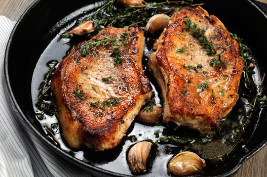 Juicy, browned pork steak on a bone in oil with garlic and herbs in a frying pan. Close-up.