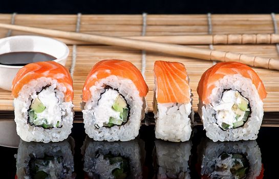 Philadelphia Sushi roll in a row. Sushi mat and sticks with soy sauce on a blurry background. Reflection.