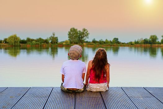Boy with flat cap and little girl are sitting on pier. Boy and girl are looking upwards on sky. Love, friendship and childhood concept. Beautiful romantic sunset picture. 