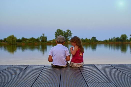 Boy with flat cap and little girl are sitting on pier. Love, friendship and childhood concept. Beautiful romantic sunset picture. Copy space.