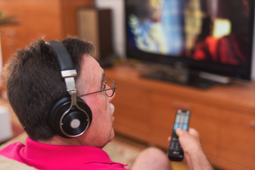 Back view of senior man with headphones watching tv holding remote control