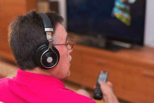 Back view of senior man with headphones watching tv holding remote control