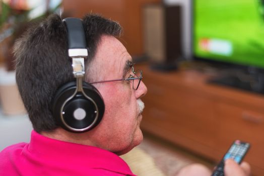 Back view of senior man with headphones watching tv holding remote control