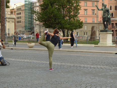 Flexible hipster girl balances in a standing pose, bending her leg back. Posing for a photo shoot in front of the monument to Julius Caesar in Rome, October 7, 2018.