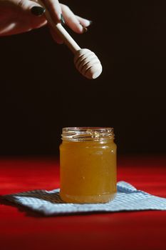 Hand with dipper picking honey from a jar of honey. Jars of honey, bee honeycomb and bee pollen on wooden table