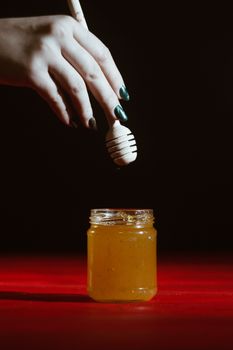 Hand with dipper picking honey from a jar of honey. Jars of honey, bee honeycomb and bee pollen on wooden table