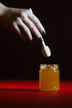 Hand with dipper picking honey from a jar of honey. Jars of honey, bee honeycomb and bee pollen on wooden table