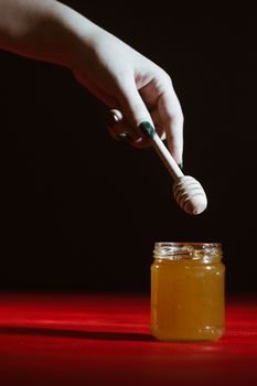 Hand with dipper picking honey from a jar of honey. Jars of honey, bee honeycomb and bee pollen on wooden table