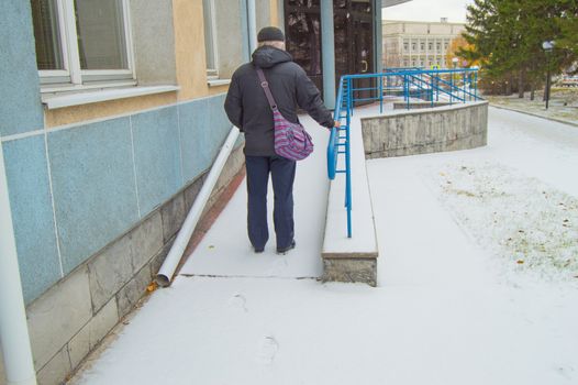An elderly gray-haired sick man climbs hard on a snow-covered ramp for the disabled, leaving traces in the snow.