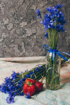 Seasonal summer flowers in vase blue cornflowers and fruits strawberries on a napkin close-up conceptual background