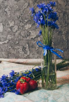 Seasonal summer flowers in vase blue cornflowers and fruits strawberries on a napkin close-up conceptual background