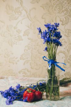 Seasonal summer flowers in vase blue cornflowers and fruits strawberries on a napkin close-up conceptual background