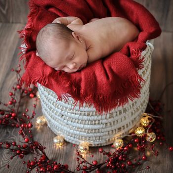 Sleeping 10 days baby on a red blanket and a white basket, surrounded by Christmas lights na dred berries.