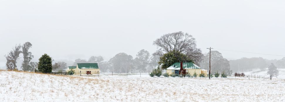 A farmhouse cream coloured with green tin roof and matching sheds and outbuildings sits in rural fields covered with fresh falling snow