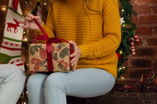 A girl in a yellow sweater unfolds a Christmas gift in a craft package. Pulling the red ribbon.