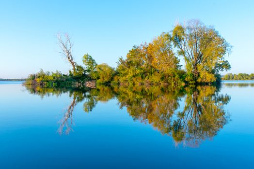 Small island with gren lush trees and clear blue sky reflected in calm water of Nove Mlyny Dam, Moravia, Czech Republic.