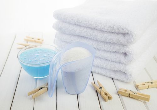 Stack of three white fluffy bath towels, washing powder in measuring cup, blue fabric softener in a transparent glass bowl and wooden clothespins on the background of white boards