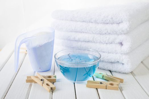Stack of three white fluffy bath towels, washing powder in measuring cup, blue fabric softener in a glass bowl and wooden clothespins on the background of white boards
