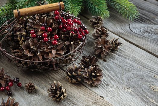 Christmas concept: full basket of pine cones and red holly berries and spruce branches on the background of old unpainted wooden boards. Christmas wallpaper