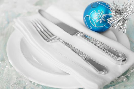 Christmas table with white plates, linen napkin, silver knife and fork and a beautiful blue Christmas ball