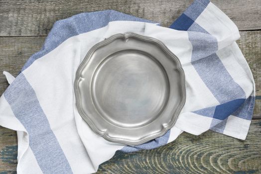 Empty pewter plate and kitchen towel on a rough wooden table, top view