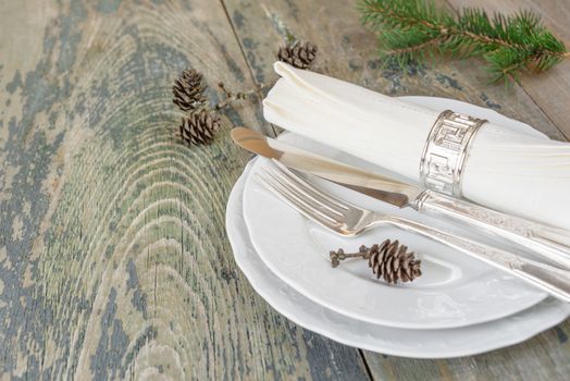 Christmas table: silver knife, fork and linen napkin lie on the white dinner plate, as well as green spruce branch and cones which is located on a old wooden table, with space for text