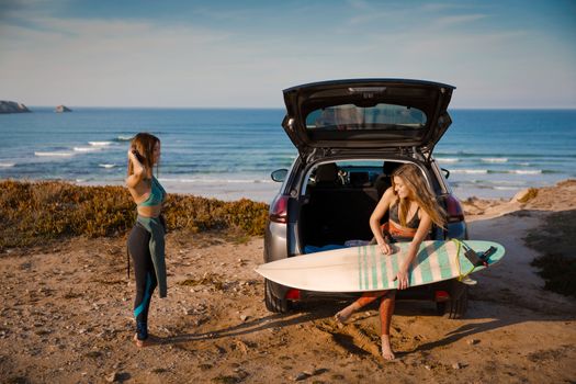 Two beautiful surfer girls near the coastline with her car, and getting ready for surfing