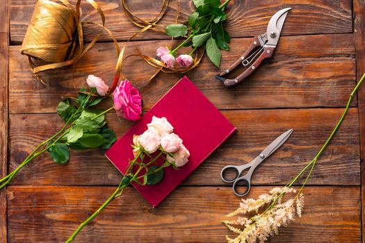 On the wooden surface laid out tools for flower bouquets: scissors, roses, colored ribbon, roses.