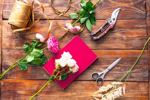 On the wooden surface laid out tools for flower bouquets: scissors, roses, colored ribbon, roses.